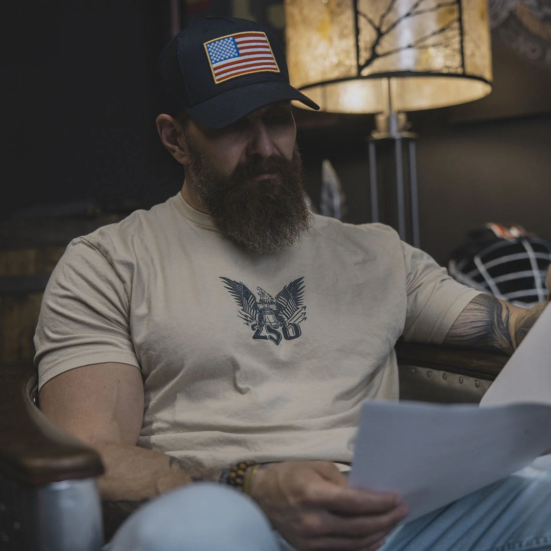 Man with a beard wearing an American flag cap and the GBRS Group Semper Fortis Short Sleeve Shirt reading a piece of paper in a dimly lit room.