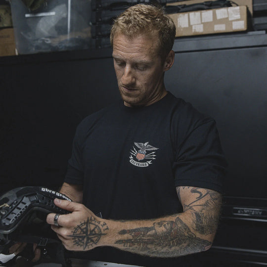 Man wearing the GBRS Group Test of Time Short Sleeve Shirt, holding a tactical helmet in a workshop setting.
