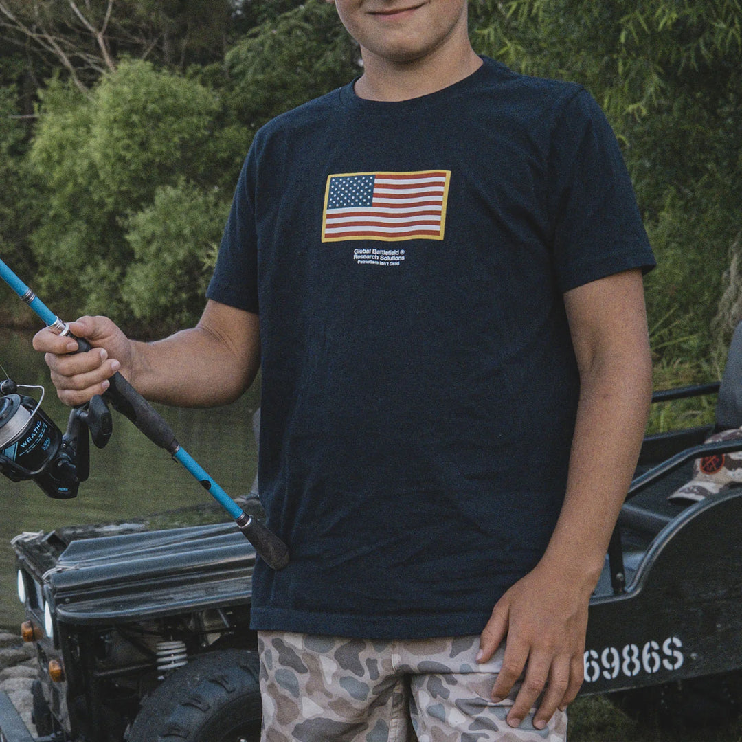 Child wearing the GBRS Group Navy Blue Youth Front & Center Short Sleeve Shirt with an American flag design, holding a fishing rod, standing next to a vehicle.
