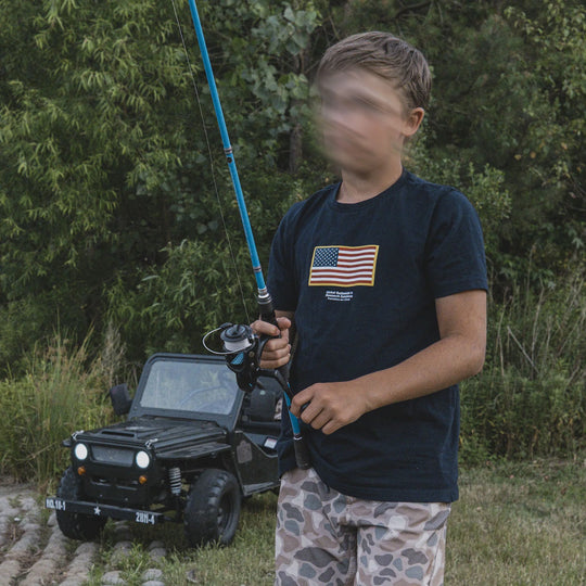 Child wearing the GBRS Group Navy Blue Youth Front & Center Short Sleeve Shirt, holding a fishing rod while standing next to a toy car in a natural setting.