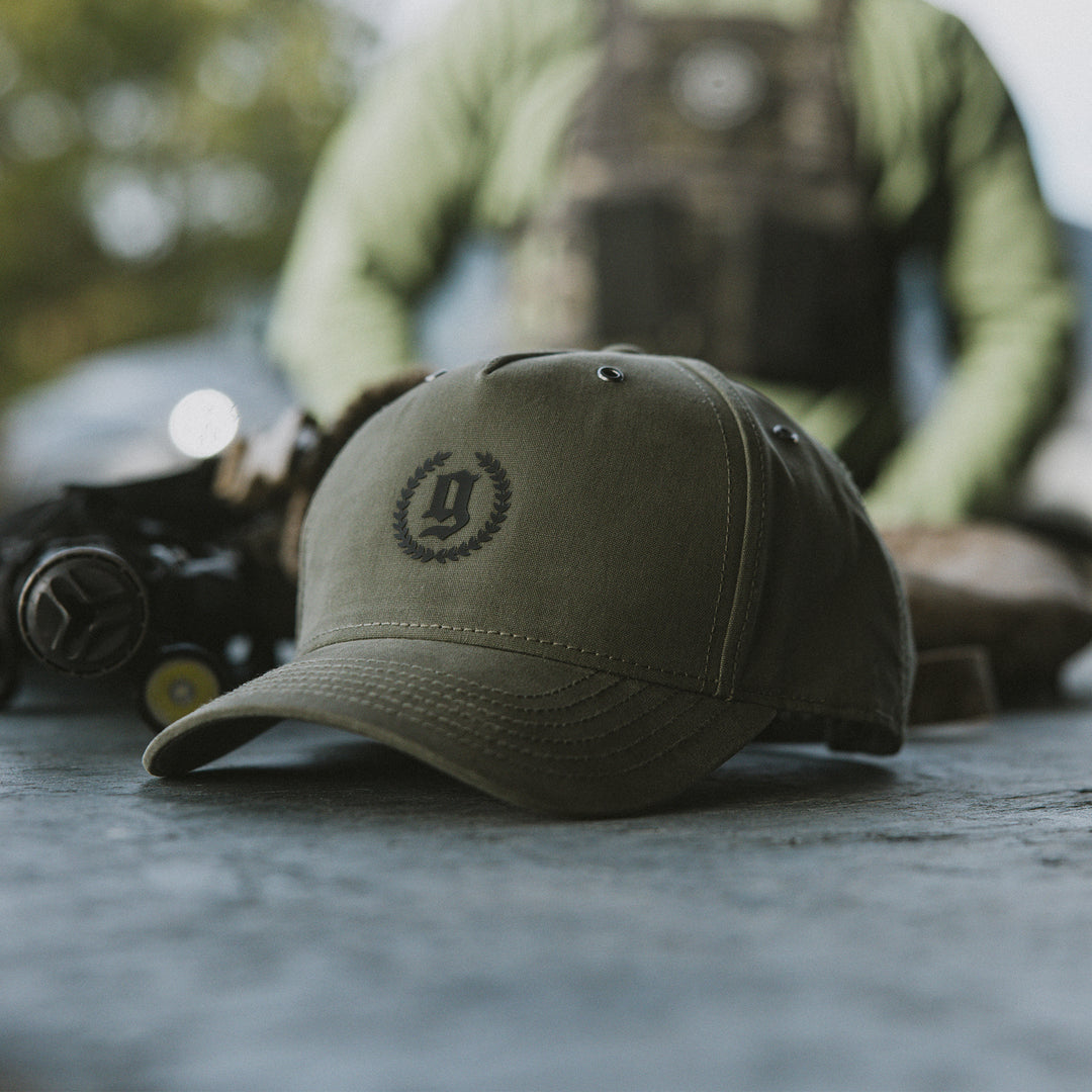 GBRS Group Legacy Canvas Trucker Hat in Loden. Front of hat pictured in lifestyle photo on table at the range.