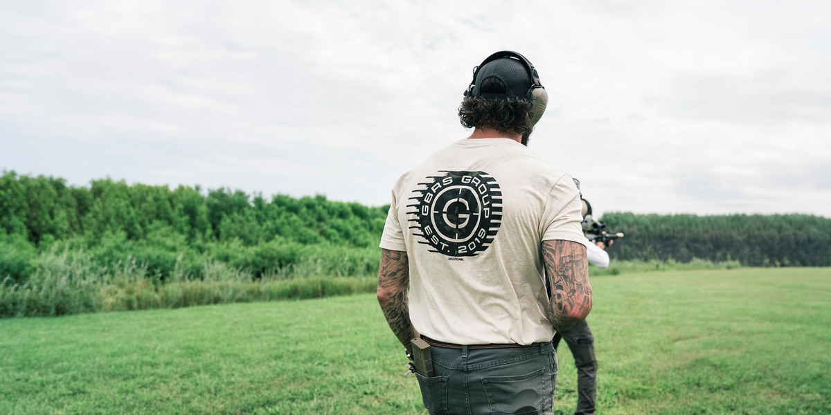 Man in a large grassy field wearing a GBRS Group tee shirt and tactical gear looking at another man aiming down a scope