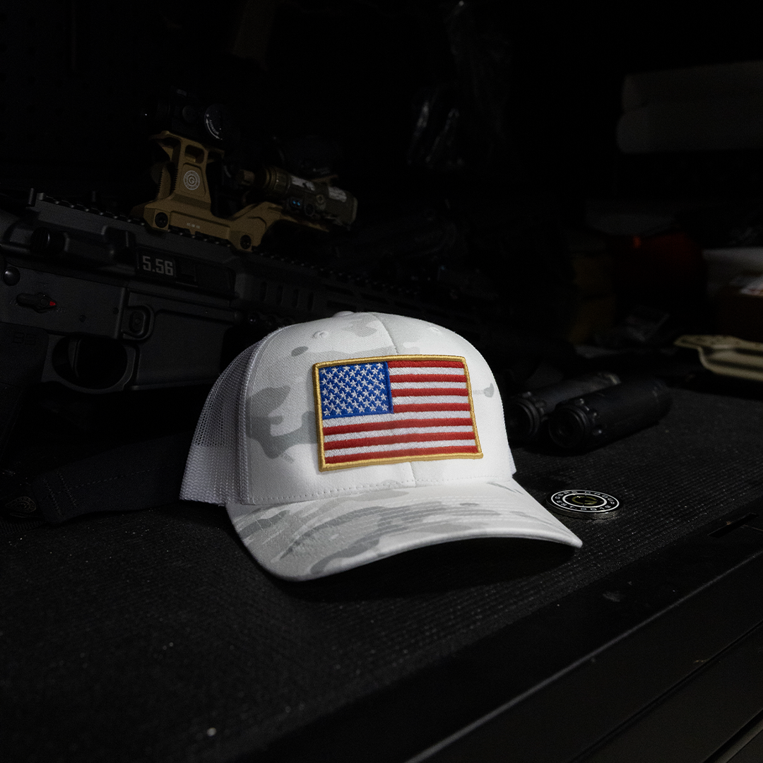 A white camouflage trucker hat with an embroidered American flag patch on the front, displayed on a dark textured surface with tactical gear in the background.