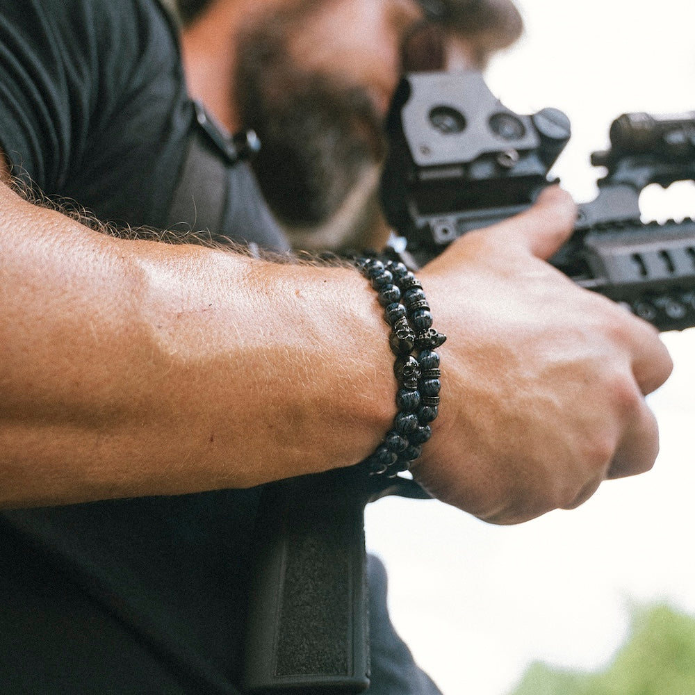 A close-up of a man's forearm and hand wearing a black beaded bracelet, holding a rifle with a red dot sight in an outdoor setting.