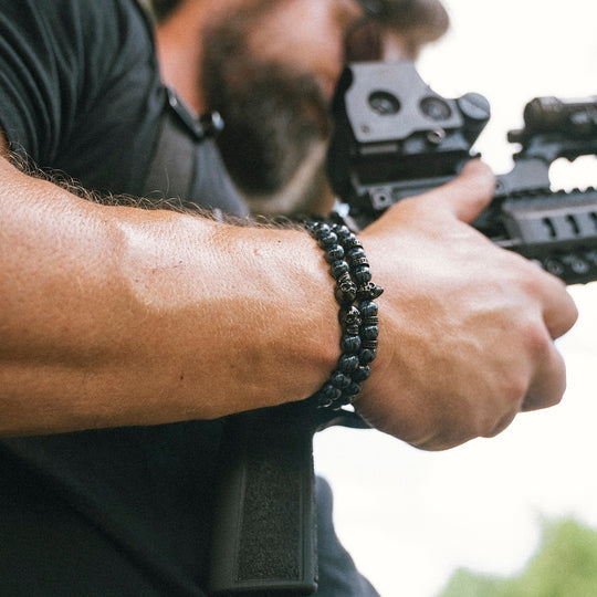 A close-up of a man's forearm and hand wearing a black beaded bracelet, holding a rifle with a red dot sight in an outdoor setting.