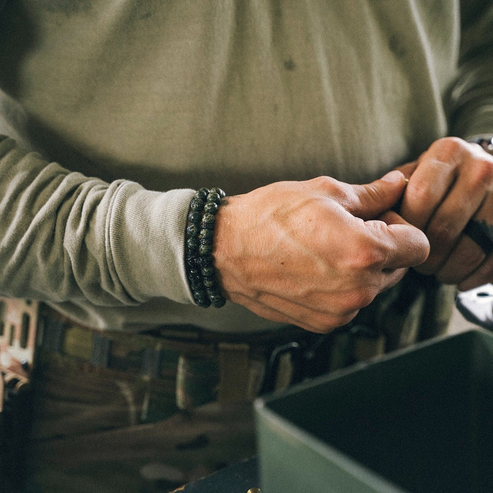 A close-up of a person's hands wearing the GBRS Group x SkeletonHD Multicam Beaded Bracelet on one wrist, wearing a muted olive green long-sleeve shirt and tactical gear.