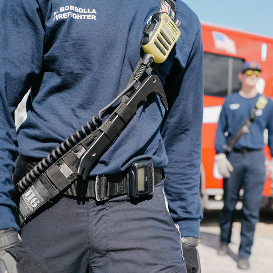 Firefighter wearing a black metal tool bar strapped to his chest, a radio, and other equipment with a fire truck in the background.