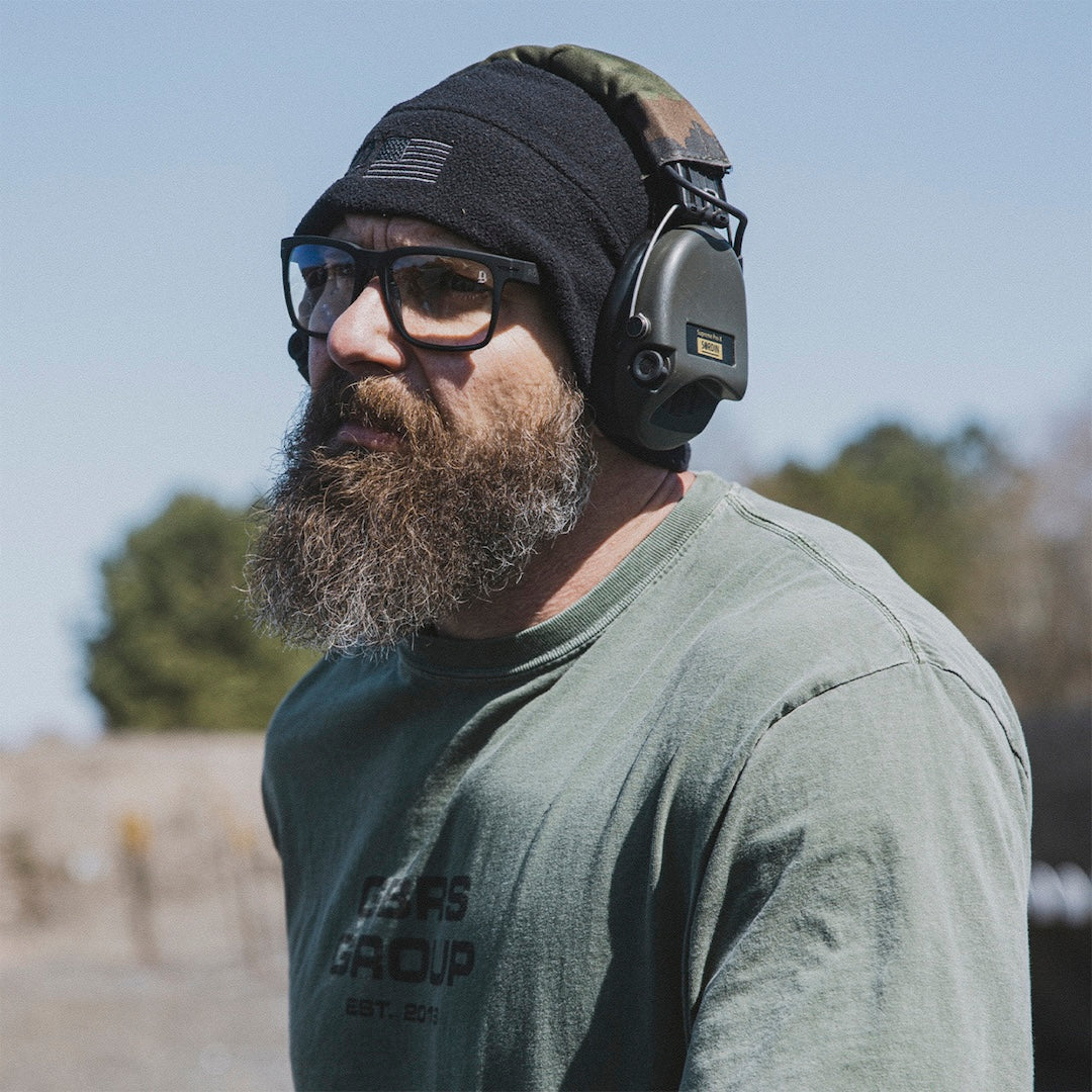 Man wearing a green shirt, black beanie with an American flag patch, ear protection, and clear glasses for eye protection in an outdoor setting. 