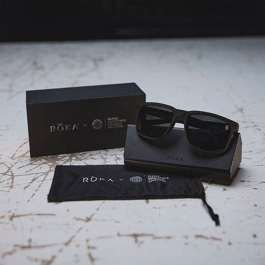 Black sunglasses with tinted lens, a case and box on a textured surface, featuring the GBRS Group and Roka brand collaboration. 